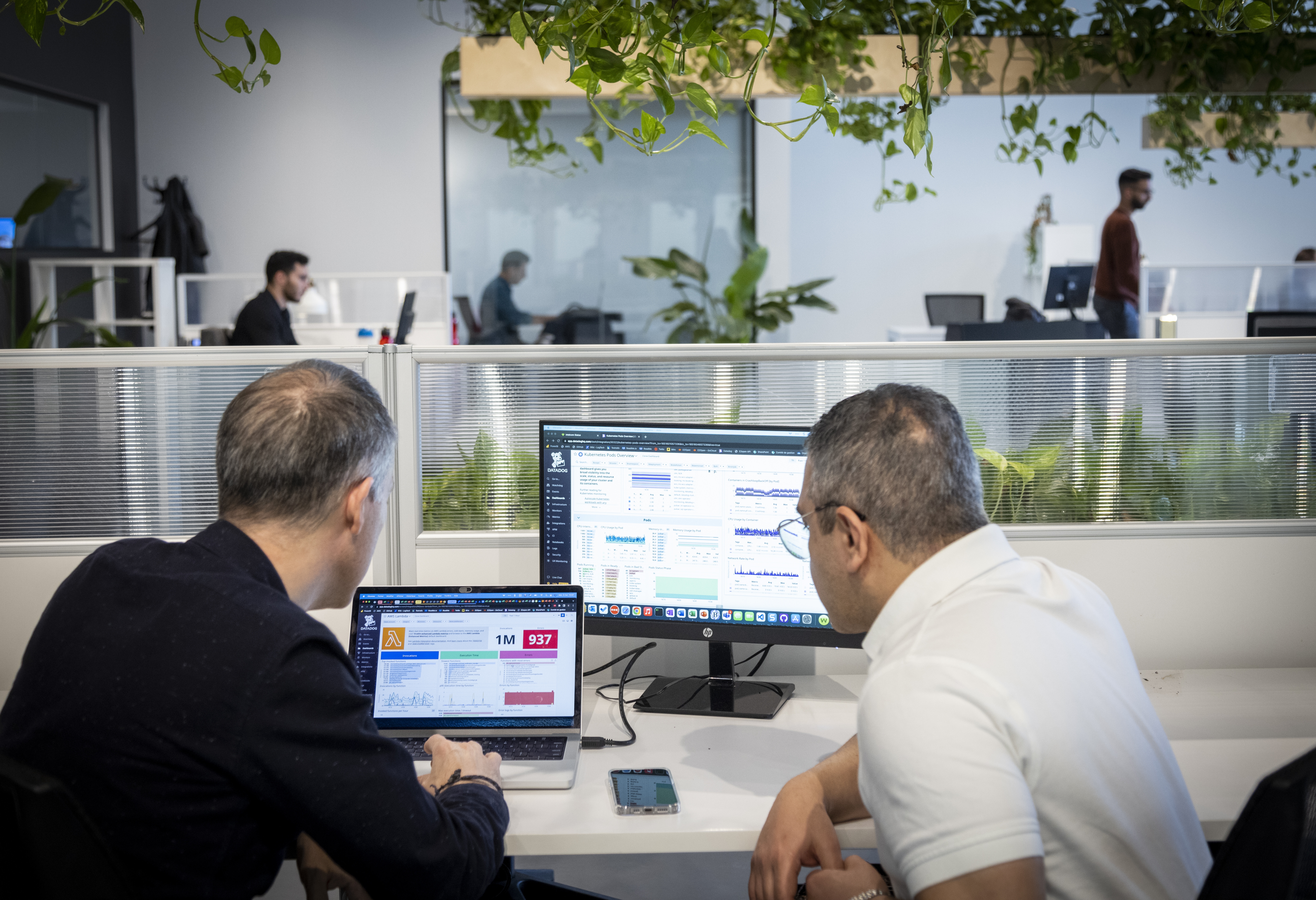 Two men are sitting at a desk and looking at two screens in an open-plan office. The background include a few more people working at computers, as well as plants.